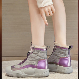 A person wearing a pair of mid-calf winter boots with quilted gray panels, metallic purple accents, and a fuzzy cream lining, shown from the knees down against a neutral indoor background.