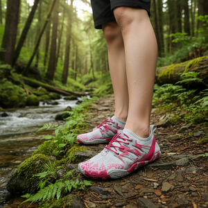 A close-up of a person's legs wearing pink and white water shoes standing on a mossy forest path next to a flowing stream.