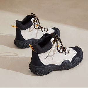 A pair of mid-top hiking boots with a white and black color-blocked design, featuring chunky black laces with gold chain details and gold heel pull tabs, set against a warm beige background.