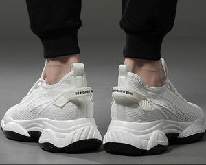 A pair of white mesh athletic sneakers with chunky white and black soles, shown from the back on a model wearing black pants against a gray studio background.