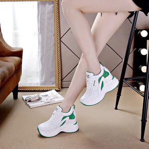 White platform sneakers with green accents worn by a model in a styled indoor setting with a mirror, brown leather chair, and black stool.