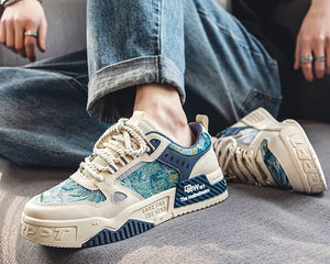 A pair of low-top sneakers shown in a styled lifestyle setting, worn with blue jeans and displayed on a gray floor. The shoes feature a cream-colored base with blue and green abstract patterned overlays, navy blue panels, and white laces.