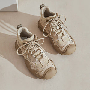 A pair of chunky low-top sneakers in a warm beige and cream colorway, featuring textured overlays, matching laces, and thick sculpted soles, photographed on a minimalist beige background with soft shadows.