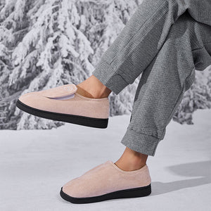 A person seated in a snowy outdoor setting wearing light blush pink slip-on slippers with black soles and gray textured pants.