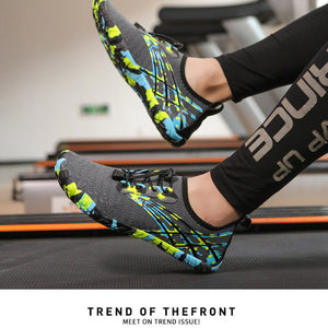 A pair of athletic sneakers with a gray mesh upper and vibrant neon green, blue, and black abstract patterns, shown being worn on a treadmill in a gym setting.