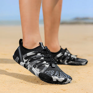 Black, gray, and white camouflage water shoes worn on a person's feet, standing on a sandy beach with the ocean and sky blurred in the background.