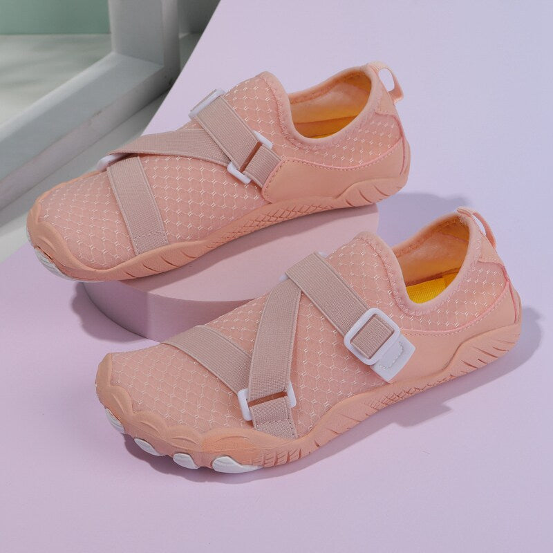 A pair of peach-colored water shoes with beige adjustable straps and white buckles, displayed on a light purple surface.
