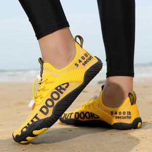 A pair of bright yellow athletic sneakers with black text reading 'OUT DOORS' and '5400 lb securin' worn on a person's feet, standing on a sandy beach with the ocean in the background.