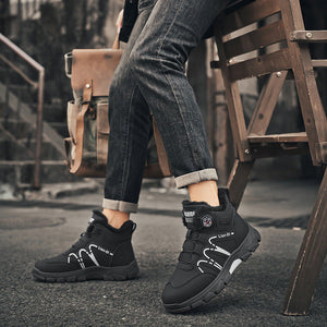 A person seated on a wooden chair outdoors wearing black mid-calf boots with white graphic details, dark jeans, and a black jacket. The background shows a rustic urban setting with stairs and pavement.