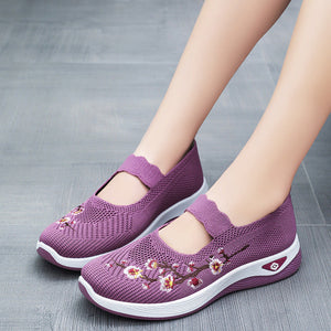 A pair of women's slip-on knit shoes in purple, featuring floral embroidery on the toe and a scalloped strap, shown on a model's feet against a gray floor background.