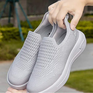 A pair of light gray slip-on mesh sneakers shown outdoors, with one shoe worn on a person's foot and the other held in a hand to show the top and side details.