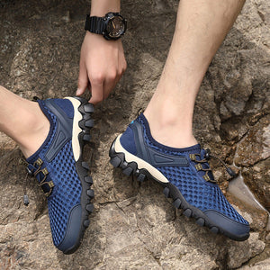 A person wearing blue mesh athletic sneakers with black and white soles, standing on a rocky outdoor surface. One hand is adjusting the strap of the left shoe, and a black wristwatch is visible on the wrist.