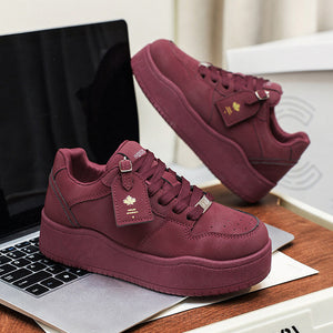 A pair of chunky low-top platform sneakers in a deep burgundy or maroon suede-like material, shown in two views: one held in a hand over a gray sofa, and another resting on a desk next to a laptop. The shoes feature matching burgundy laces, a small gold-tone logo charm, and a thick platform sole.