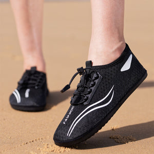 Black slip-on athletic shoes with white curved stripe accents and a visible PAXION logo, shown on a person's feet standing on sand.