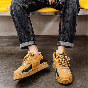 A person seated on a yellow bench wearing a brown graphic T-shirt, dark cuffed jeans, and tan-and-navy chunky sneakers in a real indoor setting.