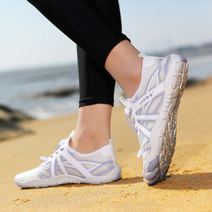 White athletic sneakers with light gray accents worn by a person standing on a sandy beach with the ocean in the background.