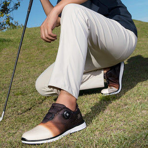 A person crouching outdoors on grass wearing a pair of golf shoes. The shoes are dark brown and beige with a gradient design and a hook-and-loop strap closure.