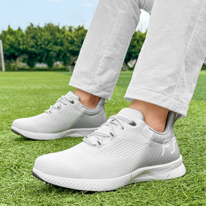 White athletic sneakers with gray accents, shown on a person standing on green grass outdoors. The person is wearing light-colored pants, and the background features a blurred green field and trees.