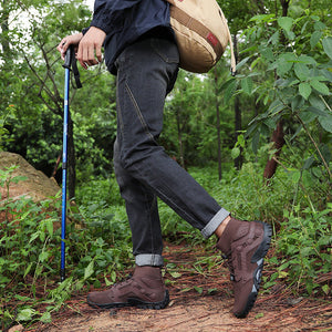 A person hiking through a green forest, wearing a navy hooded jacket, dark gray jeans, brown hiking boots, and carrying a beige backpack with a blue trekking pole.