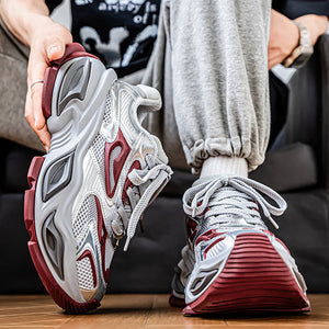 A person seated indoors wearing chunky white, gray, and burgundy athletic sneakers, with one hand lifting the left shoe to show the side and sole. Gray sweatpants and a dark shirt are visible, with a wooden floor and dark sofa in the background.