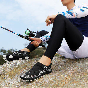 A man sitting on a rock outdoors wearing black and white patterned water shoes, black leggings, a white visor, and a blue and white long-sleeve shirt while holding a fishing rod.
