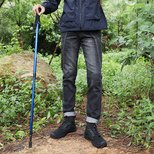 A man wearing a navy blue hooded rain jacket, dark gray jeans, and black hiking boots stands in a forest holding a blue hiking pole. He has a tan backpack on.