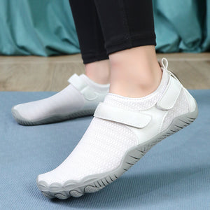 White minimalist toe-loop slip-on shoes worn on a person's feet, shown against a blue mat with a teal fabric backdrop.