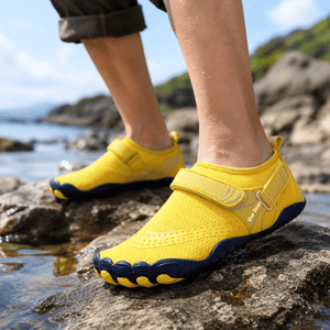 Adult wearing yellow TrailGuard wide-toe water shoes near a rocky shoreline