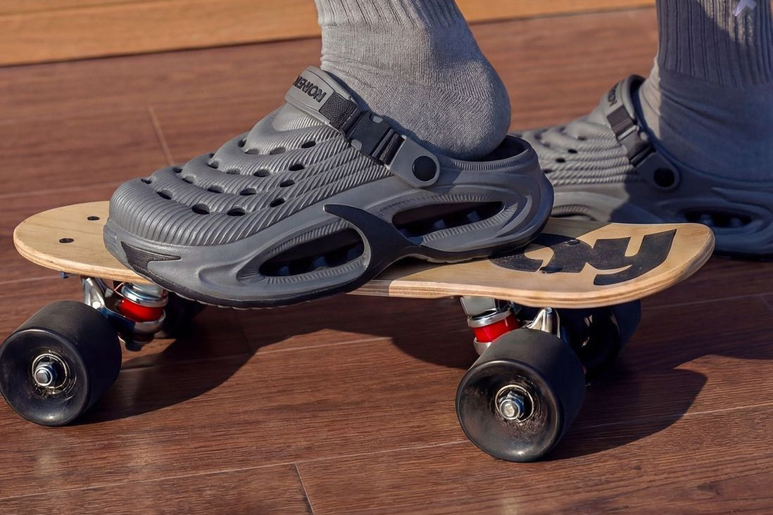 A two-panel image showing gray clog-style shoes worn with socks. The top panel shows the shoes on a wooden skateboard on a wood floor, and the bottom panel shows a close-up of a white crew sock with a black letter 'P' logo.