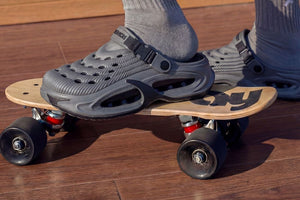 A two-panel image showing gray clog-style shoes worn with socks. The top panel shows the shoes on a wooden skateboard on a wood floor, and the bottom panel shows a close-up of a white crew sock with a black letter 'P' logo.