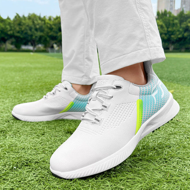 White athletic sneakers with light blue and neon green accents, worn with white pants on a green grass field with trees and buildings in the background.