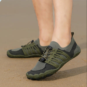 A pair of water shoes worn on a person's feet, standing on sand. The shoes are olive green and dark gray mesh with a flexible wavy sole.