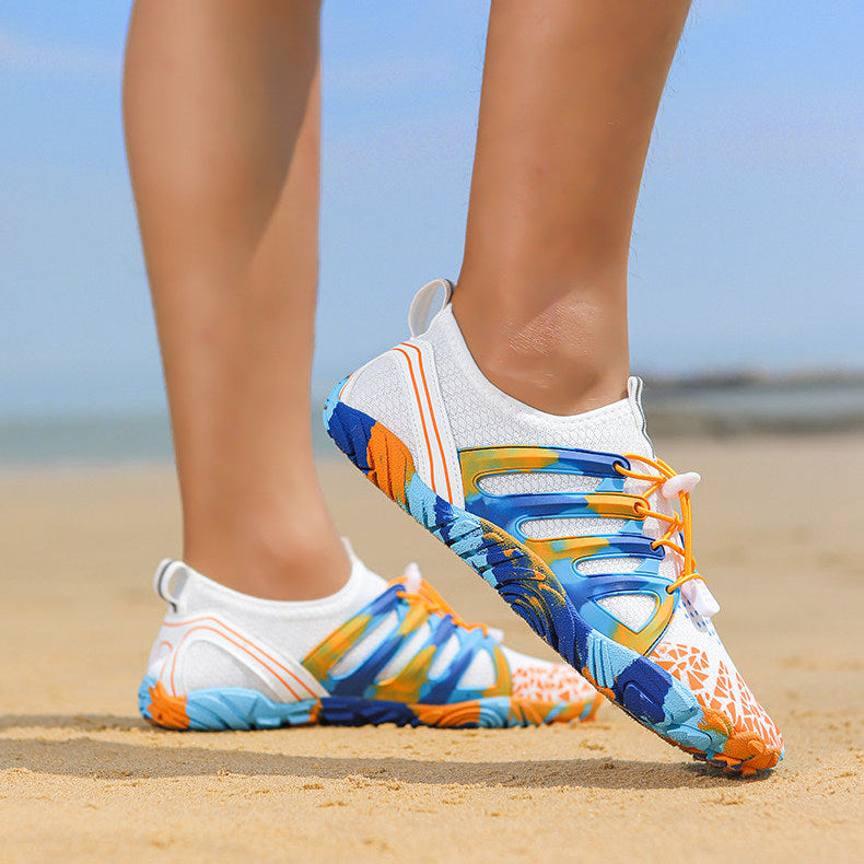 A person wearing colorful athletic water shoes on a sandy beach, with the ocean and sky blurred in the background. The shoes are primarily white with vibrant blue, orange, and yellow abstract patterns.