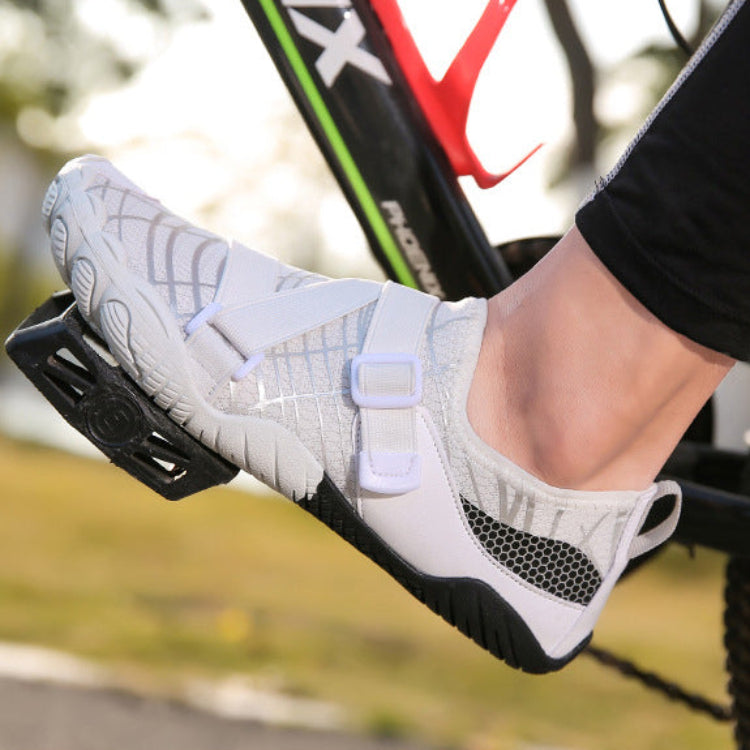 A close-up of a person's foot wearing a white and gray cycling shoe with hook-and-loop straps, mounted on a bicycle pedal.