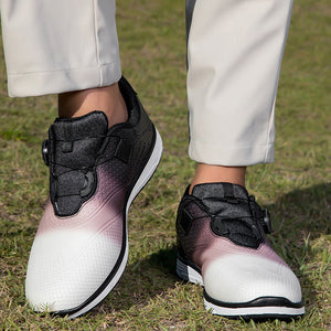A person wearing slip-on athletic shoes with a white-to-pink gradient design and black upper sections, standing on grass outdoors. The person is also wearing light beige pants.