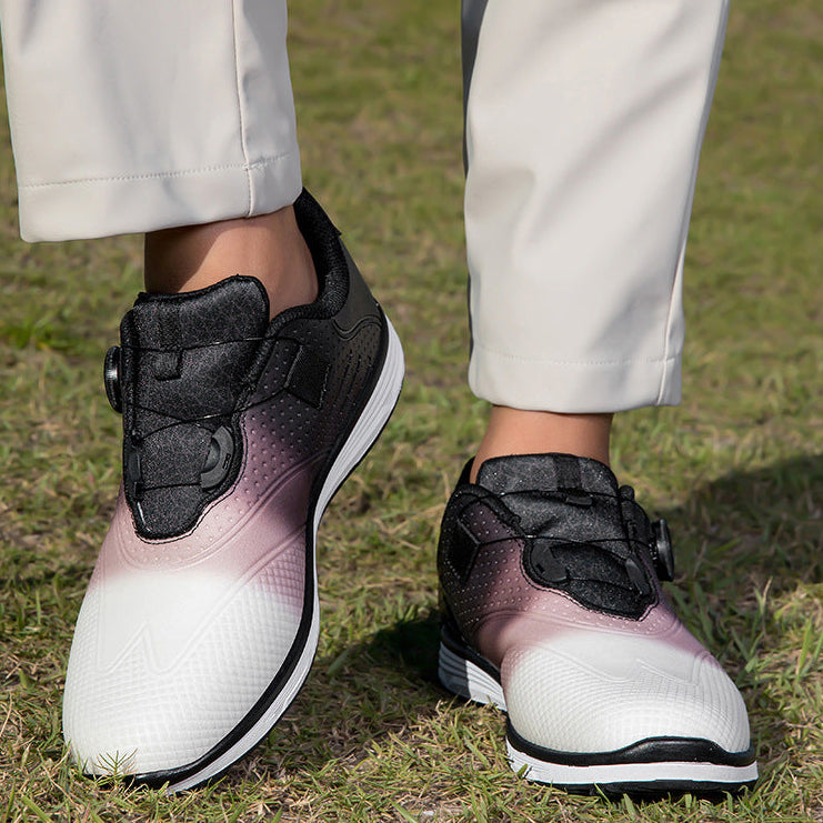 A person wearing slip-on athletic shoes with a white-to-pink gradient design and black upper sections, standing on grass outdoors. The person is also wearing light beige pants.