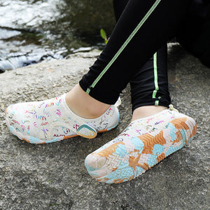 A person sitting outdoors on a stone surface near water, wearing slip-on waterproof shoes with a colorful abstract print. The person is also wearing black leggings with a light green stripe.
