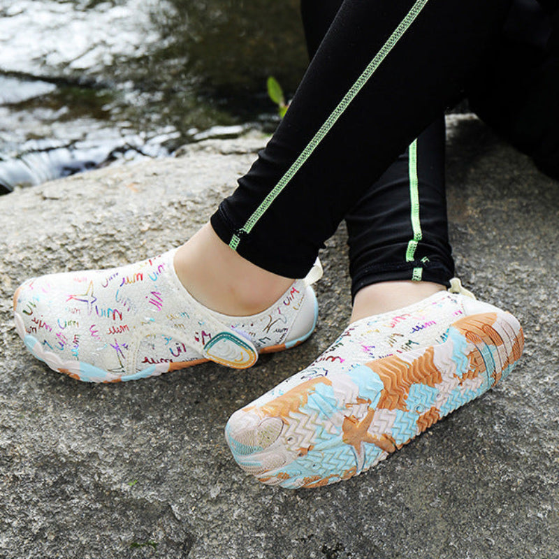A person sitting outdoors on a stone surface near water, wearing slip-on waterproof shoes with a colorful abstract print. The person is also wearing black leggings with a light green stripe.