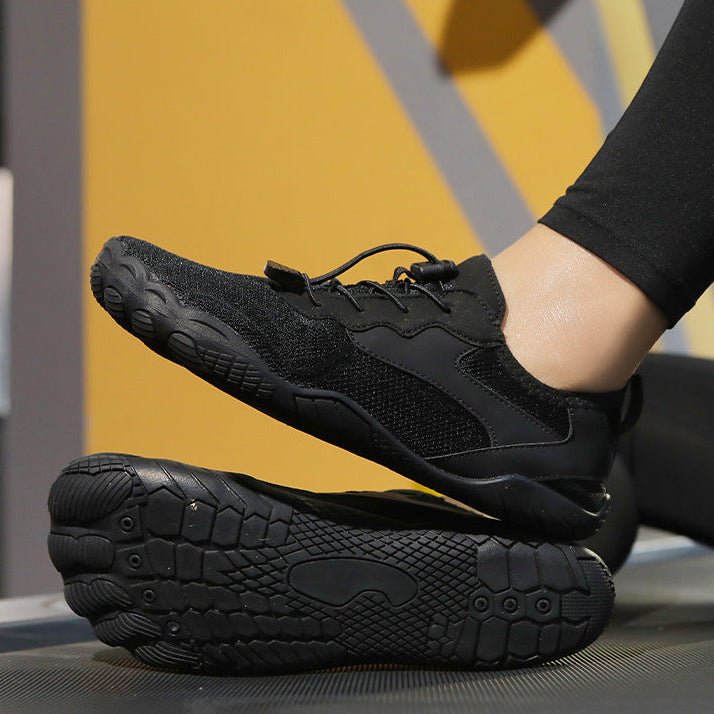 Black minimalist running shoes with a breathable mesh upper and flexible sole, shown on a treadmill with one shoe lifted by a person's foot.