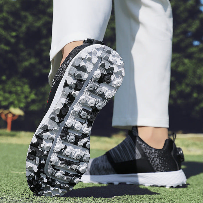 A close-up lifestyle shot of a person's legs on a golf course, showing the sole of a black and white golf shoe with black spikes, paired with white pants.