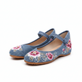 A pair of blue suede flat shoes with pink and white floral embroidery on the toe, displayed on a clean white background.
