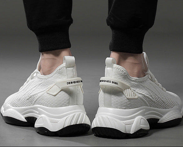 A pair of white mesh athletic sneakers with chunky white and black soles, shown from the back on a model wearing black pants against a gray studio background.