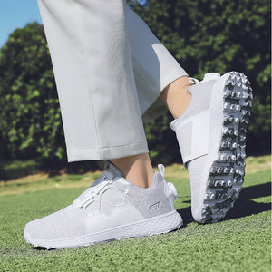 White athletic golf shoes with a textured sole, shown on a person wearing light gray pants on a green grass field with trees in the background.