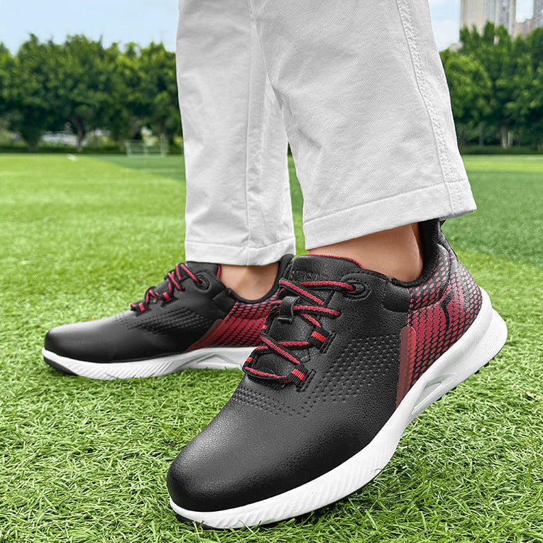A person wearing black and red athletic sneakers with white soles, standing on green grass outdoors. The photo is cropped from the knees down, showing light-colored pants and the shoes in use.