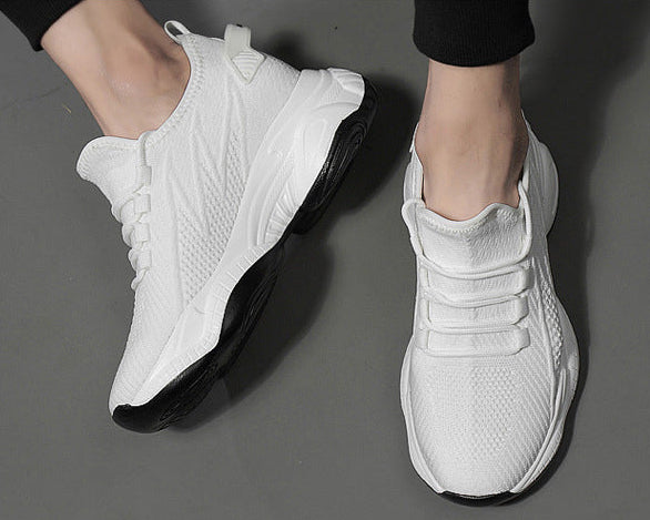 A pair of white athletic sneakers with black soles, shown on a person's feet against a dark gray floor. The top of the shoes features a textured knit-like pattern, and the sole has a chunky, sculpted design.