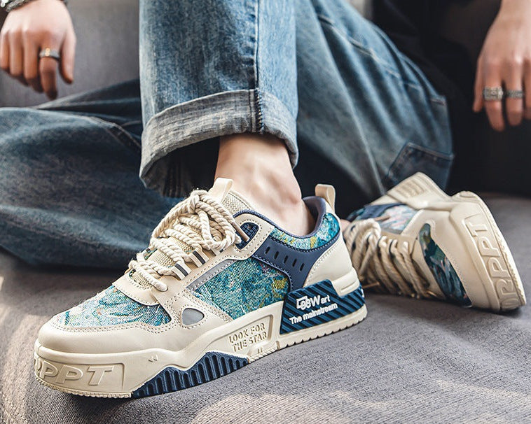 A pair of low-top sneakers shown in a styled lifestyle setting, worn with blue jeans and displayed on a gray floor. The shoes feature a cream-colored base with blue and green abstract patterned overlays, navy blue panels, and white laces.