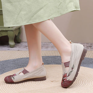 A person wearing a light sage green skirt and modeling a pair of beige slip-on mesh flat boots with burgundy floral embroidery on the toes and heels, standing on a woven straw mat.