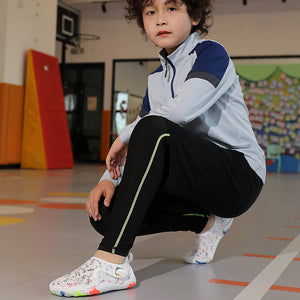 A child wearing a light blue and navy athletic zip-up jacket, black athletic pants with a green stripe, and white patterned sneakers, crouching in an indoor sports hall.
