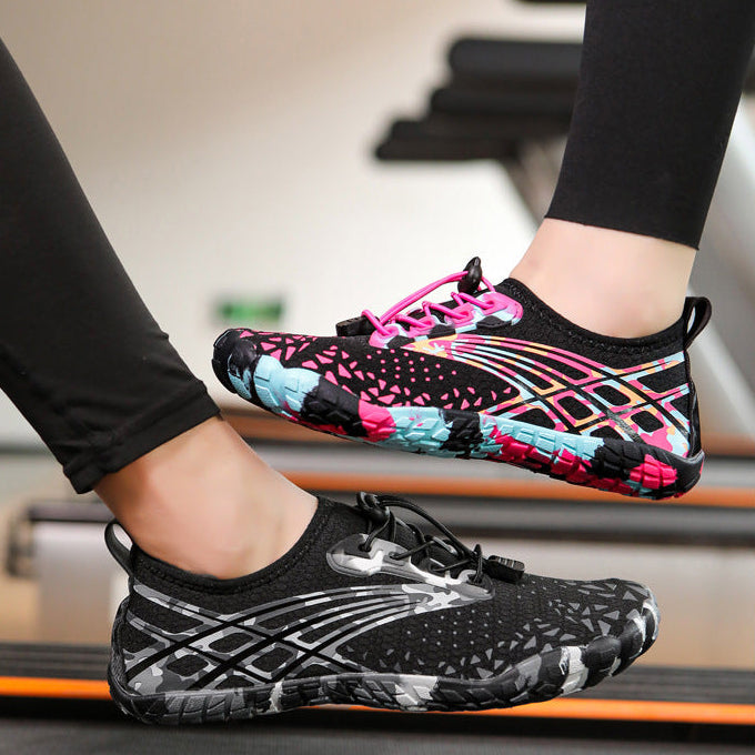 A pair of women's water shoes shown on a treadmill, with one shoe worn on a foot and the other floating above the treadmill belt. The shoes feature a black mesh upper with pink, blue, and white graphic accents.