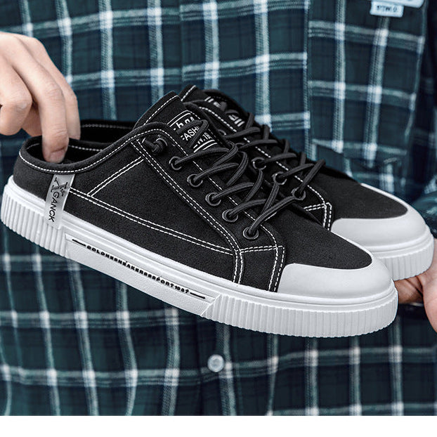 A person wearing a dark green and white plaid shirt holds up a pair of black canvas low-top sneakers with white rubber soles. The shoes feature black laces, white contrast stitching, and a small silver brand tag on the heel.